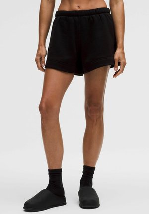 Kvinde iført sorte shorts med elastisk talje, sorte sokker og sorte slip-on sko står op ad en ensfarvet lys baggrund.