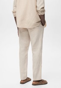 Personne portant un pantalon en lin beige et une chemise à manches longues assortie retroussées, debout les mains dans les poches et des sandales marron.