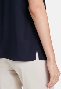 Marineblaue Bluse mit glatter Textur, seitlichen Schlitzen und lässigem Schnitt. Kombiniert mit hellen, sportlichen Hosen.
