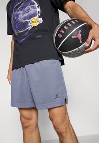Camiseta gráfica negra con diseño en púrpura y blanco, combinada con pantalones cortos perforados gris claro que presentan un logo negro de Jordan. Sosteniendo un balón de baloncesto negro.
