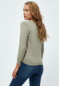 Frau mit langen, gewellten Haaren, die einen hellgrauen Pullover und blaue Jeans trägt, seitlich vor einem schlichten, hellen Hintergrund.