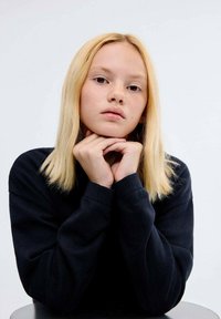 Oseba s svetlim lasem in srednje dolgimi lasmi, oblečena v temno pulover, s brado naslonjeno na prekrižane roke, proti svetli podlagi.