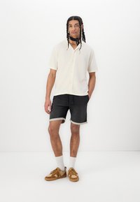 Cream short-sleeve shirt, black denim shorts with rolled cuffs, tan sneakers, white ankle socks, standing against a plain white background.