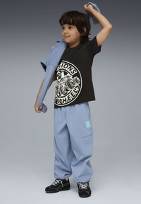 Niño con camiseta gráfica negra y pantalones azules poniéndose una chaqueta azul a juego contra un fondo gris.