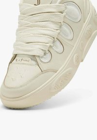 Detailní záběr krémově zbarvené tenisky s tlustými tkaničkami, síťovanými panely, raženým textem na podrážce a značkou "La Francé" na jazyku.