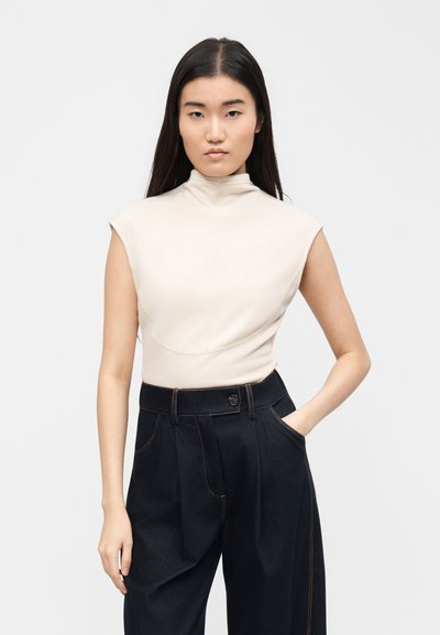 Νεαρή γυναίκα με μακριά σκούρα μαλλιά φορά αμάνικη εκρού μπλούζα με ζιβάγκο και σκουρόχρωμο παντελόνι με φαρδιά μπατζάκια και ψηλή μέση, στέκεται με το ένα χέρι στην τσέπη.