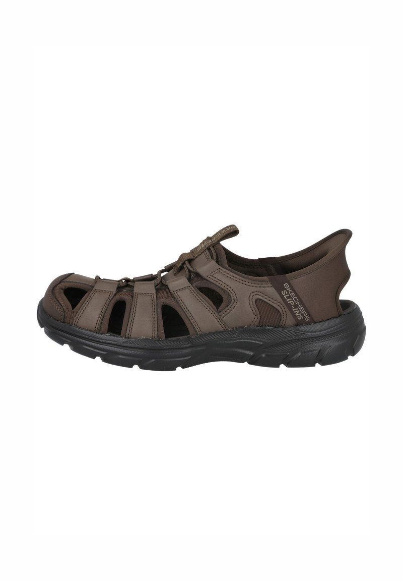 Sandaña marrón con diseño abierto, que presenta un upper texturizado, cierre elástico tipo bungee y una suela de goma negra resistente.