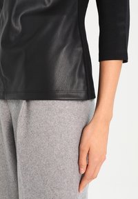 Schwarzes Lederoberteil mit figurbetontem Design, ausgestattet mit Dreiviertelärmeln und glatter Textur. Kombiniert mit hellgrauen Wollhosen.