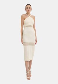 Abito aderente color crema con scollatura a collare, dettagli in crochet in vita e lunghezza al ginocchio. La modella indossa tacchi a strappo coordinati.