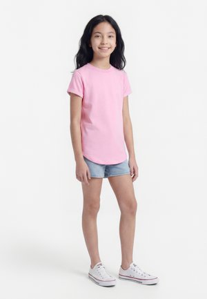 Jente som står og smiler, iført en rosa kortermet skjorte, lyse denimshorts og hvite joggesko foran en ensfarget hvit bakgrunn.