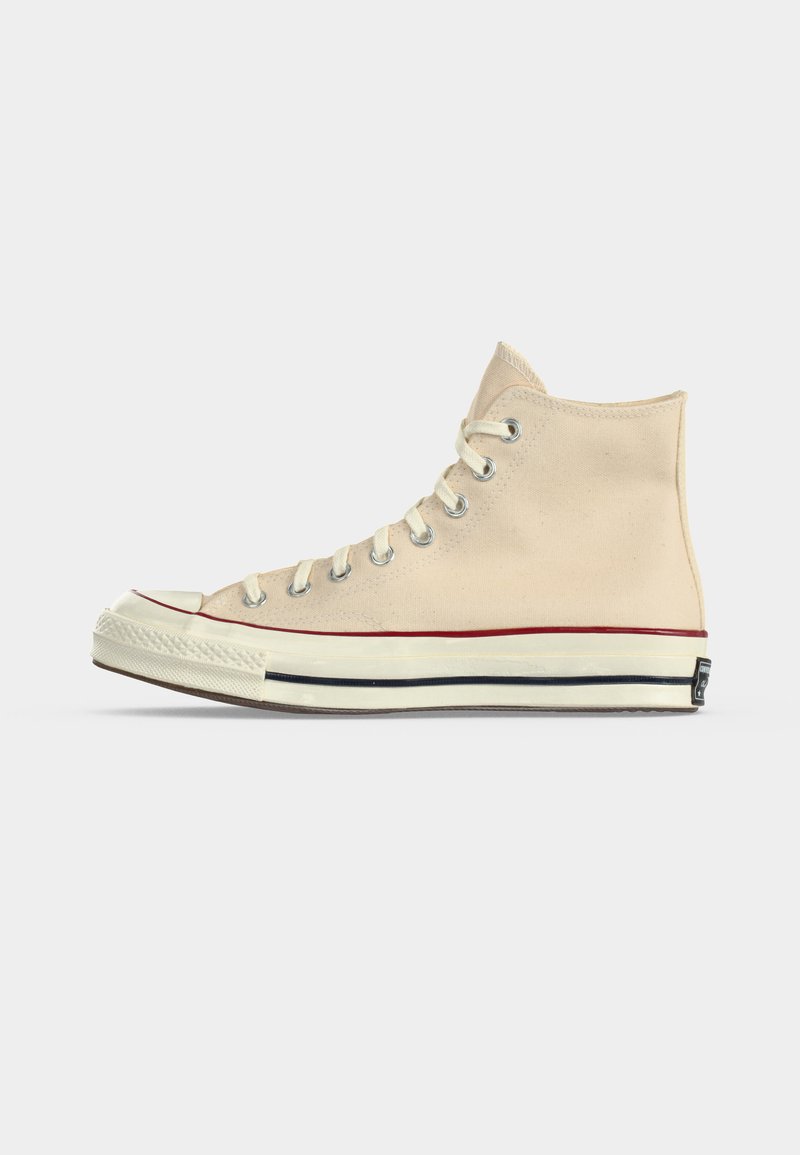 Beiger High-Top-Sneaker aus Canvas mit weißer Gummisohle, roten und schwarzen Streifen, Metallösen und weißen Schnürsenkeln, in linker Seitenansicht abgebildet.