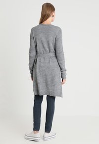 Žena stojící, oblečená v dlouhém šedém pleteném kardiganu s páskem, tmavých skinny džínách a bílých platformových teniskách, otočená zády proti jednobarevnému pozadí.