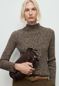 Donna bionda in un maglione lavorato a maglia marrone con motivo tiene una pochette in pelle marrone scuro, indossando una cintura beige e anelli sulle dita.