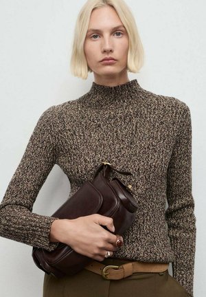Mujer rubia con un suéter de punto marrón con un patrón, sostiene un bolso de mano de piel marrón oscuro, lleva un cinturón beige y anillos en los dedos.