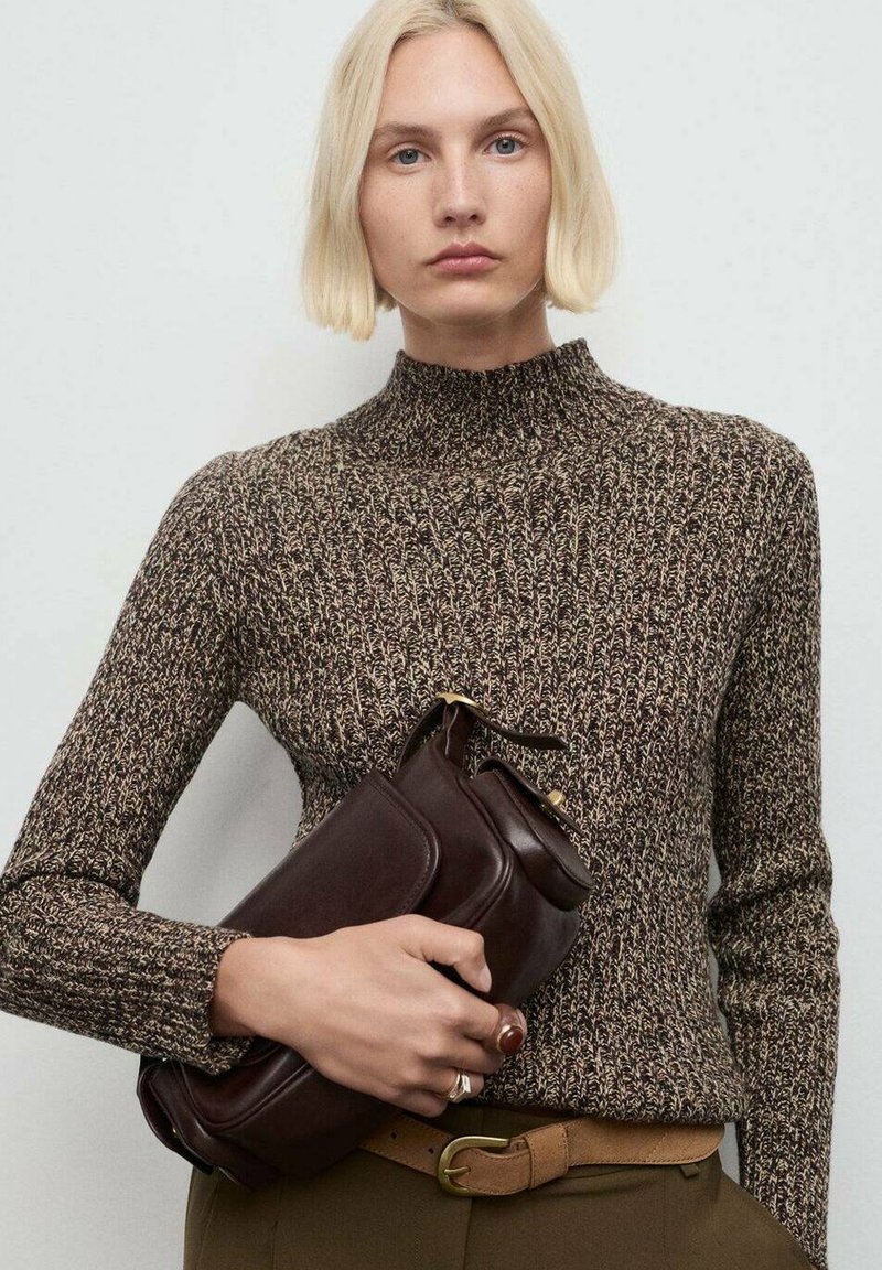 Donna bionda in un maglione lavorato a maglia marrone con motivo tiene una pochette in pelle marrone scuro, indossando una cintura beige e anelli sulle dita.