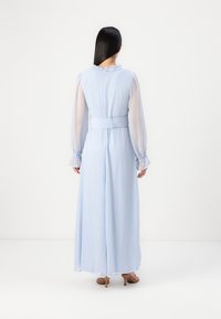 Vaaleansininen pitkä mekko, jossa on läpikuultavat hihat, röyhelökaulus ja vyötäretetty vyötärö. Materiaali vaikuttaa pehmeältä ja soljuvalta; muotoilu on elegantti ja rento siluetti.