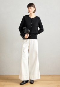 Cardigan nero a maglia con bottoni dorati, abbinato a pantaloni bianchi a gamba larga e stivaletti neri con tacco. Tiene in mano una pochette nera dalla texture lavorata.