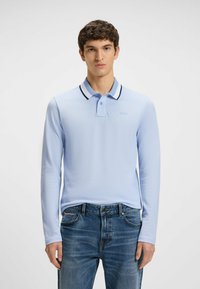 Ljusblå långärmad pikétröja med en svart- och vitrandig krage och broderad logotyp, i kombination med blå denimjeans med fickor.