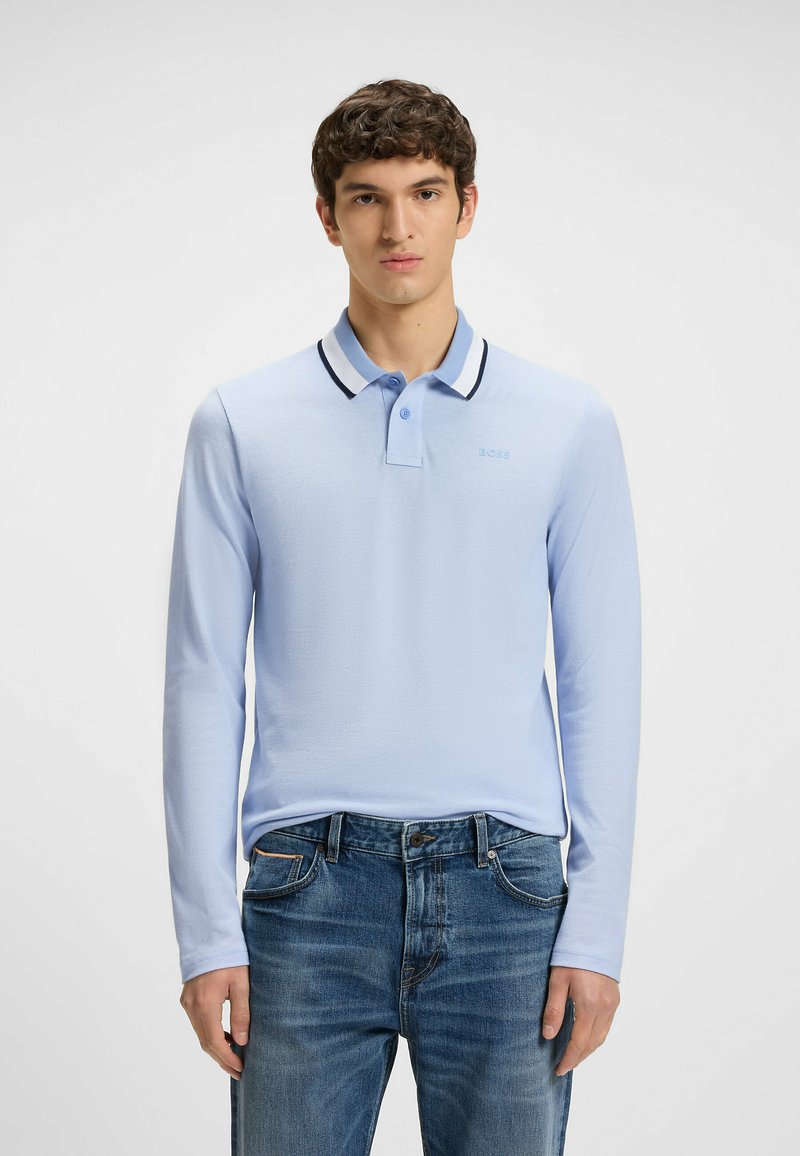 Ljusblå långärmad pikétröja med en svart- och vitrandig krage och broderad logotyp, i kombination med blå denimjeans med fickor.