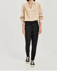 Beige Bluse mit Puffärmeln und gefaltetem Vorderteil, kombiniert mit schwarzen, taillierten Hosen mit Kordelzug und Seitentaschen, getragen mit schwarzen Turnschuhen.
