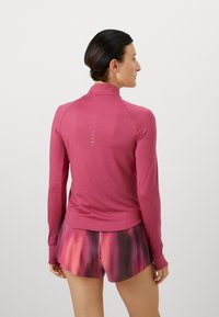 Langsleeve, nauwsluitend roze top met een hoge kraag en subtiele reflecterende details op de rug, gecombineerd met geplooide gradient shorts in rood en paars.