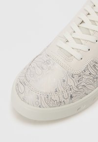 Punta di sneaker bianca con motivi astratti neri e argento testurizzati, lacci bianchi e suola in gomma su uno sfondo semplice.