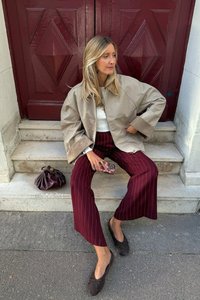 Beige bomber jacket, valkoinen toppi, maroon raidalliset housut, ruskeat matalat kengät ja ruskea käsilaukku kivisillä portailla punaisten ovien edessä.