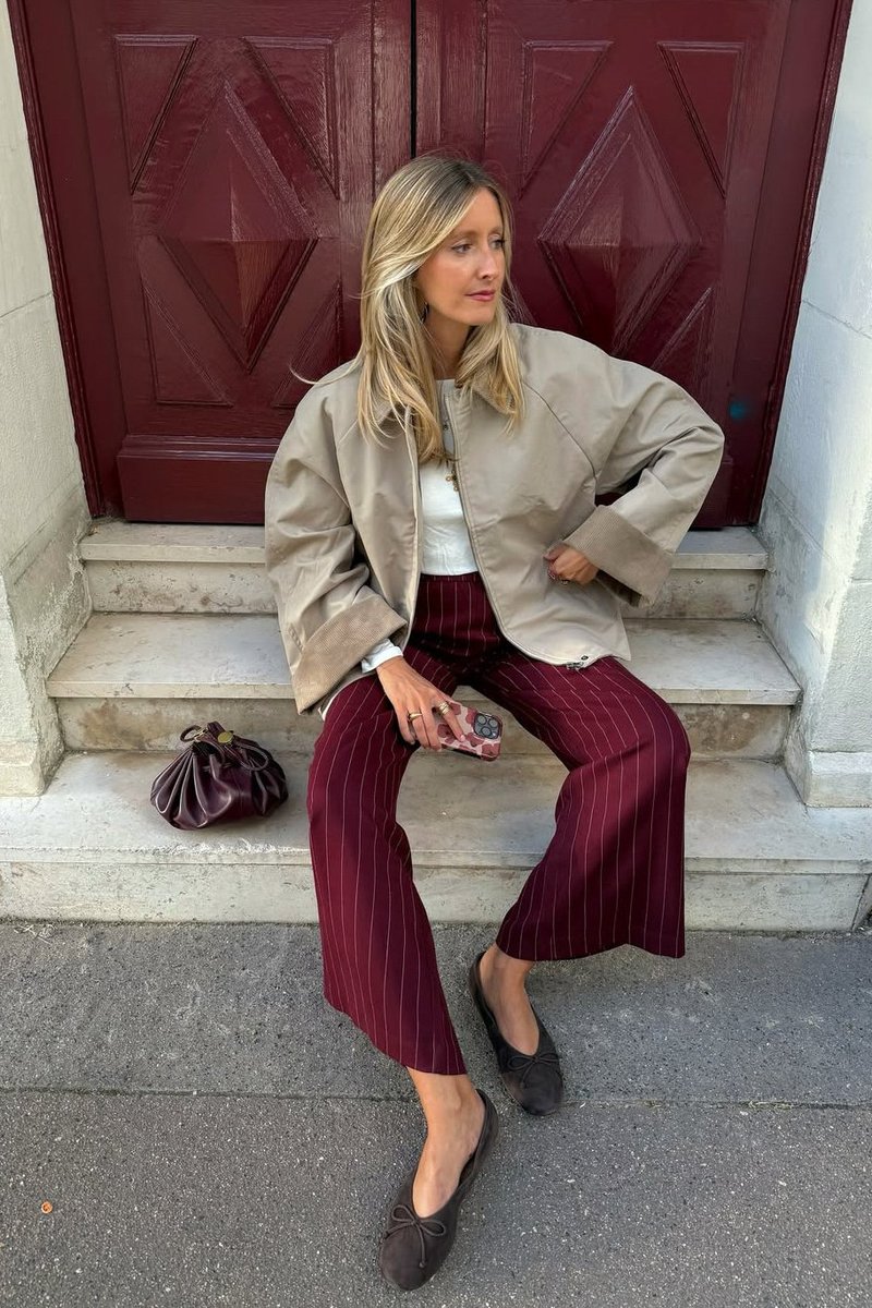 Beige bomber jacket, valkoinen toppi, maroon raidalliset housut, ruskeat matalat kengät ja ruskea käsilaukku kivisillä portailla punaisten ovien edessä.