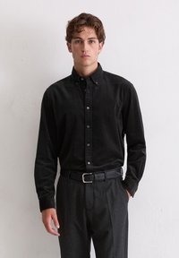 Junger Mann mit lockigem Haar, der ein schwarzes, knöpfbares Cordhemd, einen schwarzen Gürtel und dunkelgraue Hosen trägt, vor einer schlichten weißen Wand.