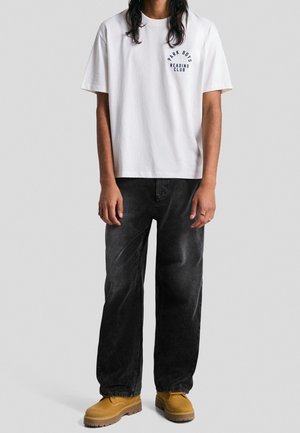 Person trägt ein weißes T-Shirt mit dem Schriftzug „Park Boys Reading Club“, dunkle weite Hose und beige Stiefel, steht vor einem schlichten grauen Hintergrund.