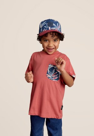T-shirt en coton rouge avec une poche bleue à motif ; casquette bleu marine avec un design de feuilles tropicales et un patch logo texturé. Jean en denim.