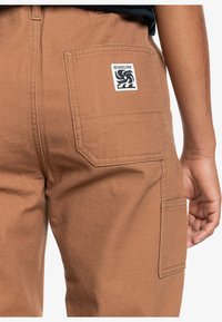 Brązowe spodnie cargo z teksturowanego materiału, posiadające tylną kieszeń z naszywką z logo oraz widocznymi detalami szwów.