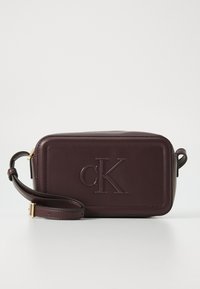 Sac en cuir bordeaux à bandoulière avec logo "cK" en relief, forme rectangulaire, bandoulière détachable et texture lisse. Accents en métal doré.
