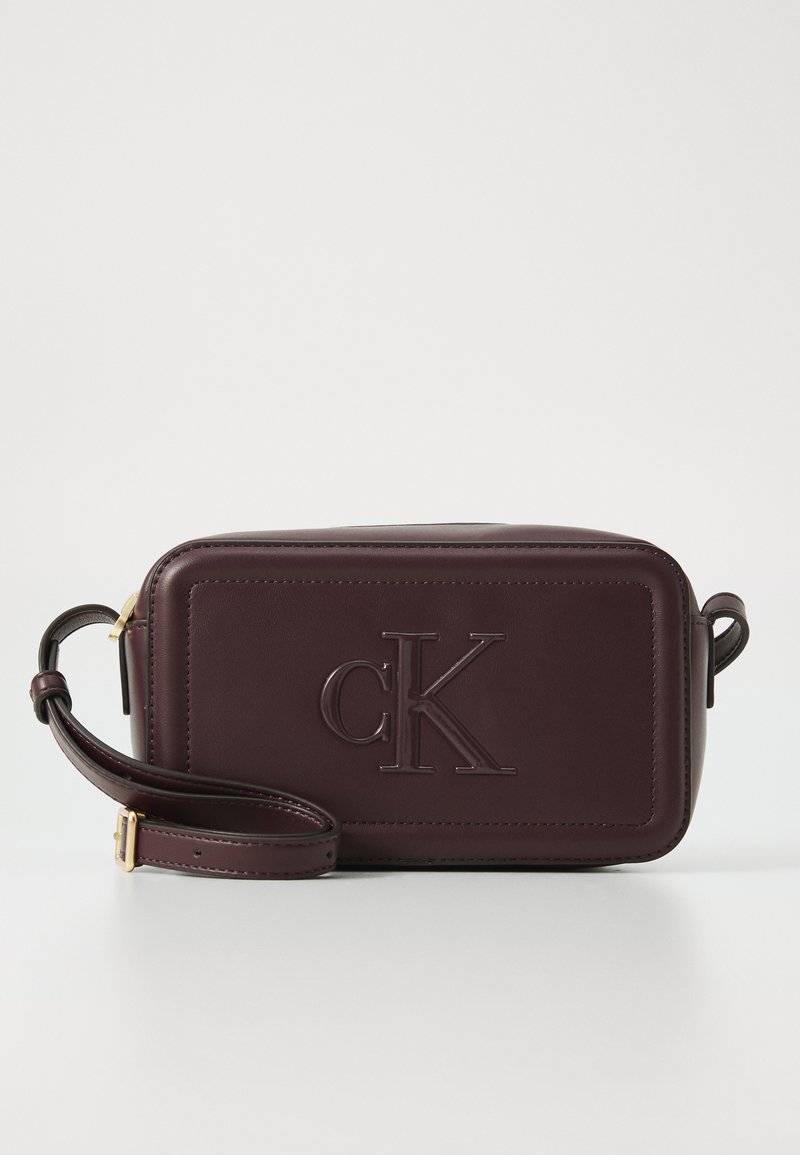Sac en cuir bordeaux à bandoulière avec logo "cK" en relief, forme rectangulaire, bandoulière détachable et texture lisse. Accents en métal doré.