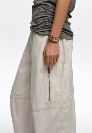 Persona que lleva pantalones anchos de pierna color beige claro con un bolsillo vertical con cremallera y un top gris fruncido a un lado, mostrando pulseras coloridas en la muñeca.