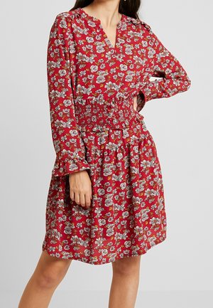 Frau in einem roten, geblümten Kleid mit langen Ärmeln, einem gerafften Taillenbereich und V-Ausschnitt, die mit einer Hand in der Hüfte steht.