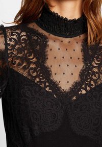 Abito nero con dettagli in pizzo, rete trasparente con pois, colletto arricciato alto e motivi floreali intricati.