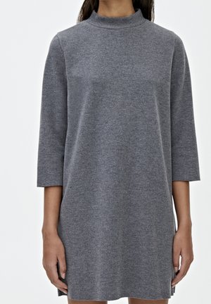 Femme portant une robe en maille gris uni à manches longues avec col montant, debout devant un fond blanc.