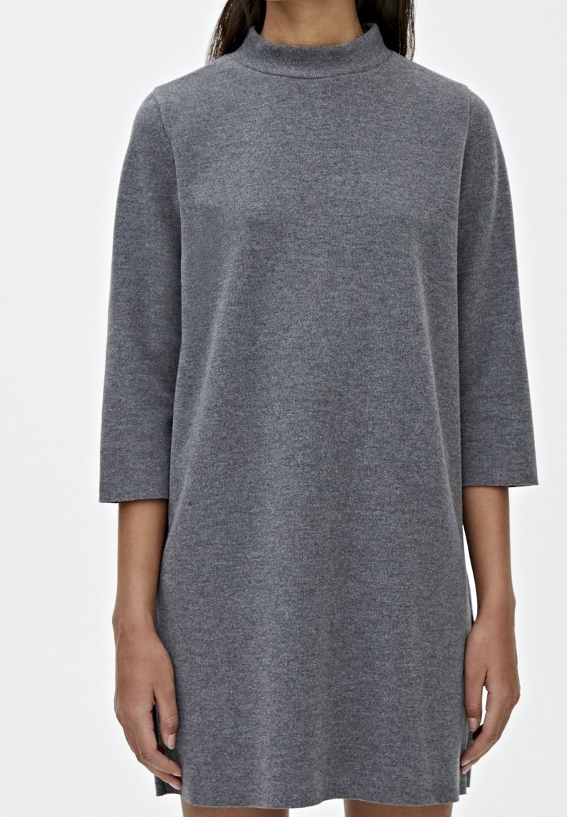 Femme portant une robe en maille gris uni à manches longues avec col montant, debout devant un fond blanc.