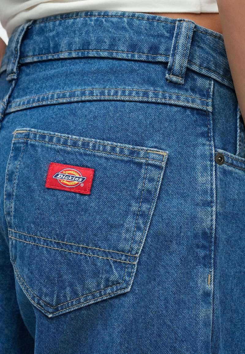 Gros plan sur une poche arrière de jean en denim bleu avec une étiquette rouge de la marque Dickies cousue dessus.