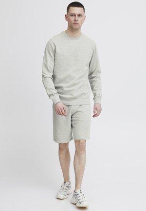 Sudadera gris claro con puños y dobladillo acanalados, acompañada de pantalones cortos a juego. Lleva unos zapatos deportivos multicolores. Diseño simple y limpio.