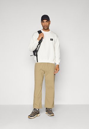 Sweatshirt blanc avec une étiquette noire, pantalon beige et baskets noires avec des accents gris. Casquette noire portée, tenant un sac à dos noir.
