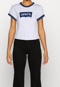 Camiseta blanca de algodón con cuello y puños azul. Presenta un logo de Levi's en color marino en el centro. Combinada con jeans negros de campana.