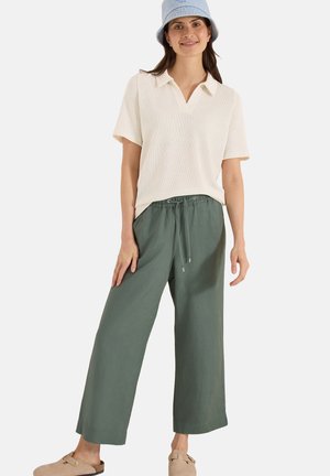 Kvinde iført en lyseblå bucket hat, cremefarvet kortærmet skjorte med krave, løse olivengrønne bukser og brune slip-on sko, stående og smilende.