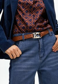 Brązowy skórzany pasek ze złotym okuciem, noszony z dżinsami i wzorzystą koszulą z pomarańczowym i granatowym wzorem. Widoczna ciemnogranatowa marynarka.