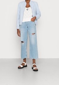 Ljust blå bomberjacka, vit stickad topp med dekorativt mönster, slitna ljusblå vida jeans och vita sandaler med svarta sulor.