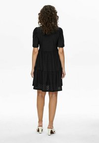 Black tiered dress with short puff sleeves, featuring a textured design and eyelet pattern; paired with white block-heeled shoes.