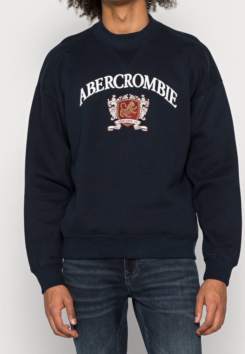 Námorně modrá mikina vyrobená z měkké látky, s nápisem "ABERCROMBIE" v bílém písmu a vyšívaným emblémem v červené a zlaté barvě. Rybímané manžety a lem.