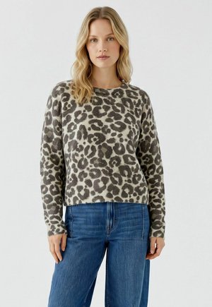Vrouw met blond haar die een grijze en zwarte luipaardprint trui en blauwe spijkerbroek draagt, staand tegen een effen lichte achtergrond.