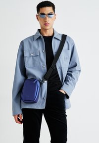 Plava lagana jakna s dva džepa na prsima, crni turtleneck, crne kordura hlače i mala ljubičasta torba preko tijela.